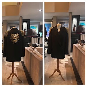 Puede incluir: Blazer negro con charreteras doradas y un diseño de cabeza de tigre dorada en la espalda. El blazer se exhibe en un maniquí de madera. La chaqueta tiene un diseño clásico con un toque moderno, adecuado para ocasiones formales o semiformales.