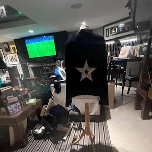 Op de afbeelding: Zwarte jas met een grote, glinsterende ster op de rug, tentoongesteld op een houten standaard. De achtergrond omvat een televisie, kunstwerken en meubels.