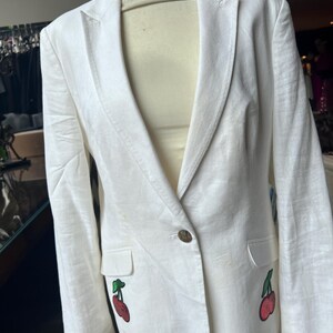 May include: A white linen blazer with a single gold button closure. The blazer has two embroidered cherry designs on the front.