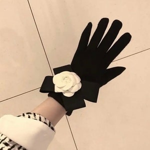 Op de afbeelding: Zwarte handschoenen met een witte bloemaccent. De handschoenen hebben een strikdetail bij de pols en een witte bloem in het midden van de strik. De handschoenen zijn gemaakt van een zacht materiaal.