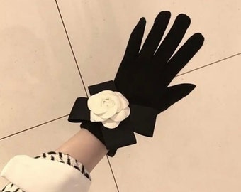 May include: Black gloves with a white flower accent. The gloves have a bow detail at the wrist and a white flower in the center of the bow. The gloves are made of a soft material.