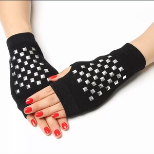 Puede incluir: Un par de guantes sin dedos negros con tachuelas plateadas en forma de pirámide. Los guantes están hechos de un tejido suave y de punto.