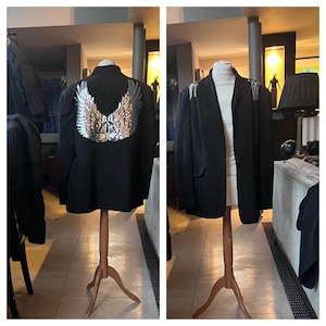 Può includere: Un blazer nero con ali d'angelo in paillettes argentate sulla schiena. Il blazer è esposto su un manichino di legno. La parte anteriore del blazer presenta abbellimenti sulle spalle. Stile classico.