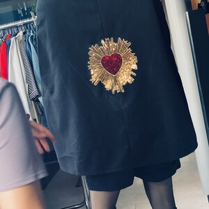 May include: A black cape with a gold heart-shaped embellishment with red glitter. The heart is surrounded by a gold sunburst design.