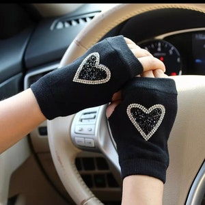 May include: A pair of black fingerless gloves with a rhinestone heart design. The gloves are being worn by someone driving a car.