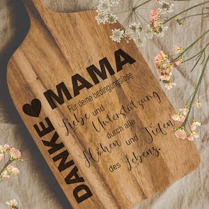 May include: A wooden cutting board with a heart and the text "DANKE MAMA" engraved on it. The text continues with "Für deine bedingungslose Liebe und Unterstützung durch alle Höhen und Tiefen des Lebens."