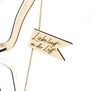 May include: A wooden flag with the text "Liebe liegt in der Luft" hanging from a string. The flag is made of light wood and has a dark brown border.