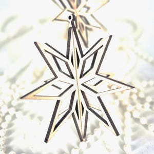 Puede incluir: Adornos navideños de madera con forma de estrella y diseño geométrico. Los adornos son de madera clara con bordes oscuros y cuelgan sobre una superficie blanca. La estrella tiene un agujero en la parte superior para colgar.