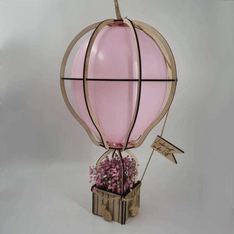 May include: A wooden hot air balloon with a pink balloon inside and a basket filled with dried flowers. The balloon has a sign that says "Liebesluft in der Luft".