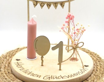 May include: Wooden birthday cake topper with a pink candle, a balloon, and the number one. The topper also has a banner that says "Jonte" and the words "Herzlichen Glückwunsch" which means "Happy Birthday" in German.