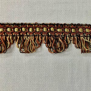 May include: Close-up of decorative trim with a repeating pattern. The trim features a woven top section in gold, brown, and black, with a fringe of dangling threads in similar colors. The background is a plain, off-white fabric.