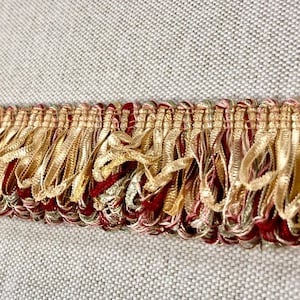 May include: A decorative fringe trim featuring a horizontal woven header with a repeating pattern of metallic gold, deep red, and muted green threads. The lower edge consists of dense, looped gold ribbon strands that create a textured effect.