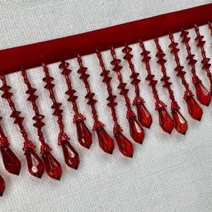 May include: Red beaded fringe trim with teardrop-shaped beads. The fringe is attached to a red ribbon.
