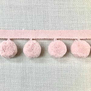May include: A close-up of a pink pom-pom trim. The trim features a woven band with a scalloped edge, from which soft, round pom-poms are suspended. The background is a light, textured fabric, providing a neutral backdrop for the decorative trim.