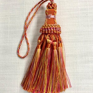 May include: A multi-colored tassel with a long, thin cord. The tassel is made of orange, yellow, pink, and white threads. The tassel is tied at the top with a knot.