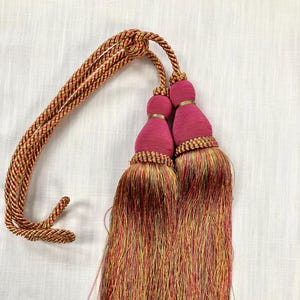 May include: A pair of pink tassel tiebacks with gold accents and a multi-colored, striped cord.