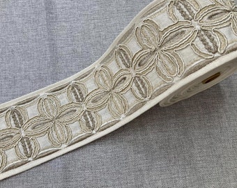 May include: A beige fabric trim with a repeating pattern of brown embroidered flowers. The trim is rolled up and ready to be used for sewing or crafting projects.
