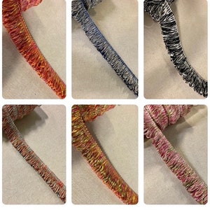 May include: A collection of decorative fringe trim in various colors. The trims include shades of red, blue, black, and multi-colored options. Each trim has a textured, fringed edge, suitable for embellishing garments or crafts.