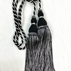 May include: Black and white decorative tassel pair. The tassels have a black and white twisted cord and a black bulbous top with a silver accent. The bottom of the tassels are black and white strands.