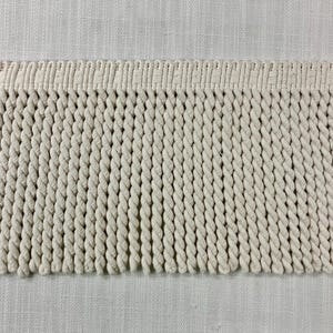 以下が含まれることがあります： ベージュの綿フリンジトリムのクローズアップ。フリンジはねじれたコードで作られており、幅は約2.5cmです。