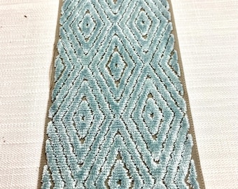 May include: Close-up of a decorative trim with a repeating diamond pattern. The trim features a soft, textured, light blue velvet design with a brown border. The trim is placed on a white fabric background.