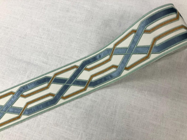 May include: A white fabric trim with a blue and gold geometric pattern. The pattern is made up of interlacing lines that create a series of squares and rectangles.