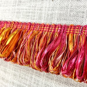 May include: Close-up of a pink and orange fringe trim. The fringe is made of a soft, silky material and has a subtle shimmer.