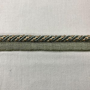 May include: Close-up of decorative trim featuring a braided rope design in shades of green and tan, with a solid green fabric base. The trim is set against a neutral, textured background.