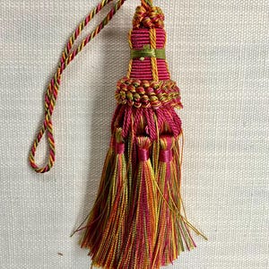 May include: A pink, green, and gold tassel with a long cord. The tassel is made of multiple strands of yarn and has a decorative knot at the top.