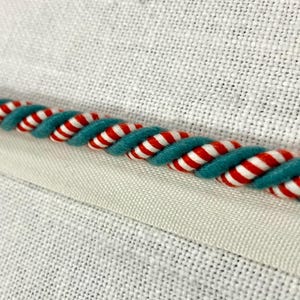 May include: Close-up of a teal and white striped rope trim with red stripes. The trim is sewn onto a white fabric.