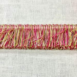May include: Pink and gold fringe trim with a woven texture. The fringe is made of multiple strands of yarn in a variety of colors.
