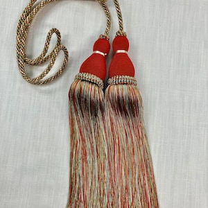 May include: A pair of red tassel tiebacks with multi-colored fringe. The tiebacks are made of a soft, silky material and have a gold cord.