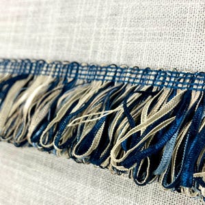 May include: Close-up of a blue and beige fringe trim. The fringe is made of a woven fabric with a subtle texture. The trim is perfect for adding a touch of elegance to any project.