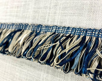 May include: Close-up of a blue and beige fringe trim. The fringe is made of a woven fabric with a subtle texture. The trim is perfect for adding a touch of elegance to any project.