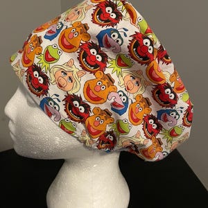 May include: A white scrub cap featuring a repeating pattern of various Muppet characters including Kermit the Frog, Miss Piggy, Fozzie Bear, Animal, and Gonzo. The fabric is printed with colourful illustrations of these iconic figures across the entire surface.