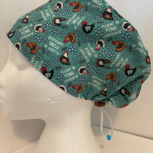 May include: A teal blue scrub hat with a white drawstring and a blue bead. The hat has a repeating pattern of chickens and flowers with the text "Fluent in Fowl Language" throughout.