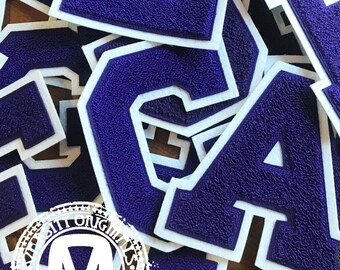May include: A selection of purple chenille letters with white felt edging. The letters are of varying sizes and orientations, arranged in a scattered pattern. The letters have a textured finish, and the overall look is reminiscent of varsity or school spirit items.