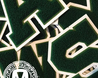 Könnte beinhalten: Eine Ansammlung von dunkelgrünen Varsity-Buchstaben-Aufnähern mit einer strukturierten Chenille-Oberfläche und einem weißen Filzrand. Die Buchstaben liegen übereinander, wobei der Buchstabe A inmitten anderer Zeichen hervorsticht.