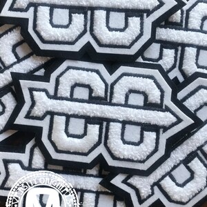 Pode incluir: Patches de letras universitárias brancas e pretas com uma textura difusa. Os patches apresentam o número "88" em uma fonte arrojada e estilizada com uma contorno preto. Os patches são provavelmente para jaquetas ou outras roupas.