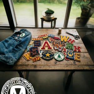 May include: A collection of varsity patches and a denim jacket are arranged on a wooden table. The patches feature various colors, letters, and designs, including a tiger, a star, and the words "Varsity" and "Tigers".