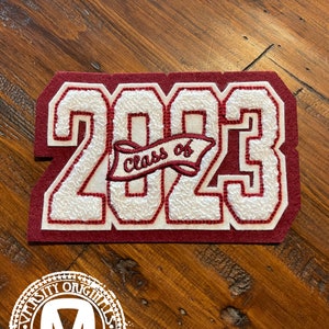 Op de afbeelding: Een bordeauxrode en witte vilten patch met de tekst "2023" in grote, getextureerde cijfers. Een banner met de tekst "Class of" is in het midden geborduurd. De patch is rechthoekig en heeft een gestikte rand.