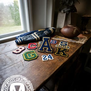 Pode incluir: Uma cole&ccedil;&atilde;o de patches de letras universit&aacute;rias em v&aacute;rias cores, incluindo azul, vermelho, verde e dourado, dispostos em uma superf&iacute;cie de madeira. Uma jaqueta universit&aacute;ria vintage azul e dourada tamb&eacute;m est&aacute; presente. Os patches apresentam letras e n&uacute;meros.