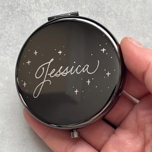 Könnte beinhalten: Ein schwarzer Kompaktspiegel mit dem Namen "Jessica" in geschwungener Schrift, umgeben von kleinen Sternen. Der Spiegel hat eine glänzende Oberfläche und einen Klappverschluss. Der Kompaktspiegel wird in einer Hand gehalten.