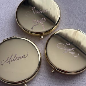 Puede incluir: Tres espejos compactos redondos de color dorado. Cada espejo tiene un nombre grabado en cursiva. Un espejo tiene el nombre "Susan" con un diseño de lazo. Los otros espejos tienen los nombres "Milena" y "Sofia".