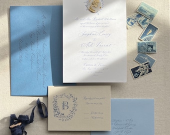 Puede incluir: Conjunto de papelería de boda elegante con sobres, invitación y tarjeta de confirmación en azul claro. La invitación tiene caligrafía y un sello de cera dorado. Sellos vintage y carrete de cinta azul.