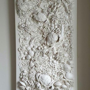 May include: A white rectangular relief sculpture depicting marine life. The artwork features detailed textures and forms, including seashells, coral, and other sea elements, creating a three-dimensional effect. The piece is mounted on a white background.
