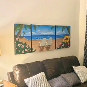 May include: A three-panel canvas wall art depicting a beach scene with palm trees, white beach chairs, and a sign that reads "Bar Beach". The canvas is mounted on a white wall above a brown leather couch with blue and white patterned pillows.