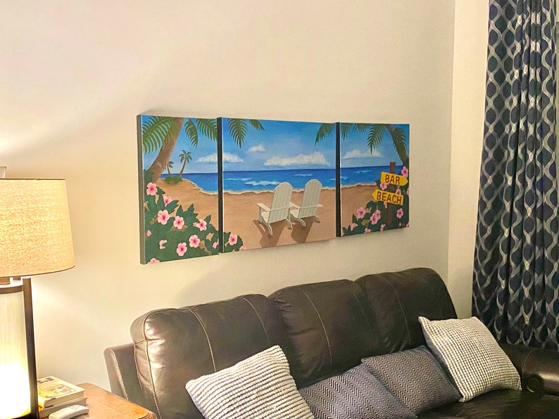 May include: A three-panel canvas wall art depicting a beach scene with palm trees, white beach chairs, and a sign that reads "Bar Beach". The canvas is mounted on a white wall above a brown leather couch with blue and white patterned pillows.