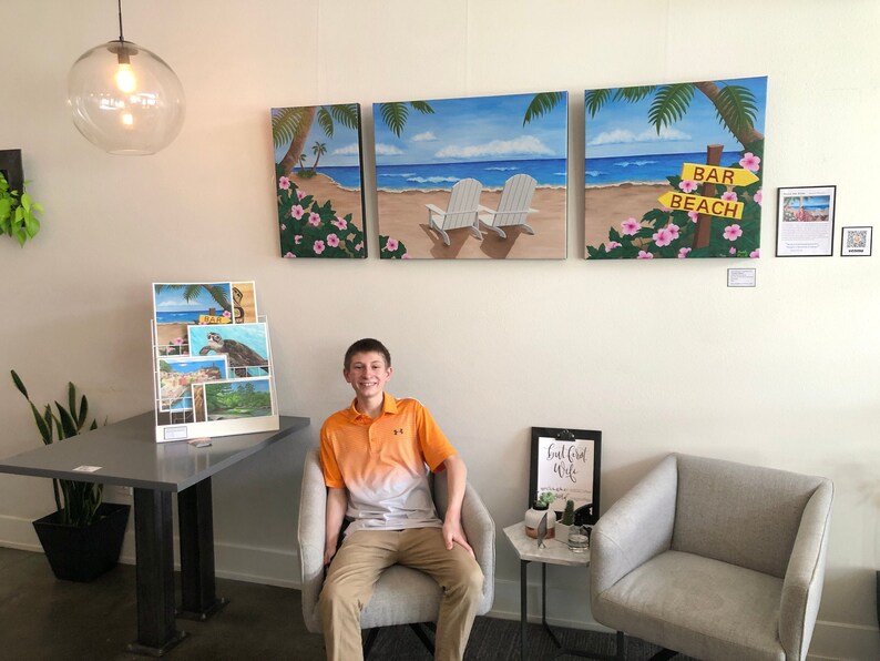 May include: A young man sits in a chair in front of three paintings depicting a beach scene with palm trees, white chairs, and a sign that reads "Bar Beach".