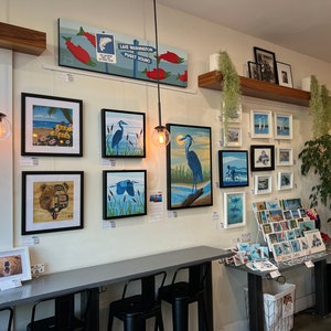 K&ouml;nnte beinhalten: Eine Galeriewand mit gerahmten Kunstwerken, darunter Gem&auml;lde von V&ouml;geln, Landschaften und einem B&auml;ren. Die Kunstwerke sind an einer wei&szlig;en Wand mit einem Holzregal dar&uuml;ber angebracht. Auf einem Tisch im Vordergrund befinden sich auch einige Postkarten und Gru&szlig;karten.